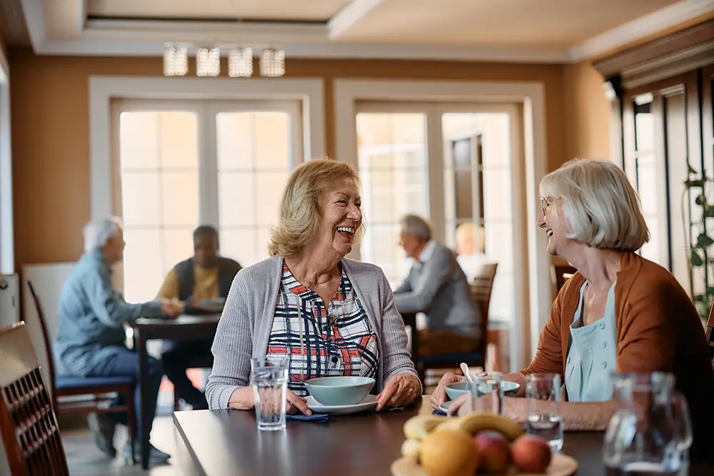 Two older women sit at a dining table, smiling and talking, with other seniors conversing in the background in a bright, communal space.