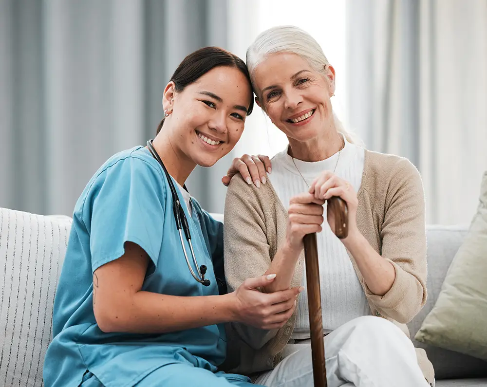 A nurse in blue scrubs sits beside an older woman holding a cane; both are smiling and sitting on a couch.