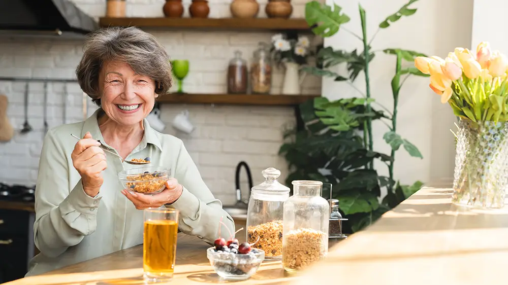 An older woman sits at a kitchen counter, smiling while eating granola with fruit. A glass of juice and jars of cereal are on the counter beside her.