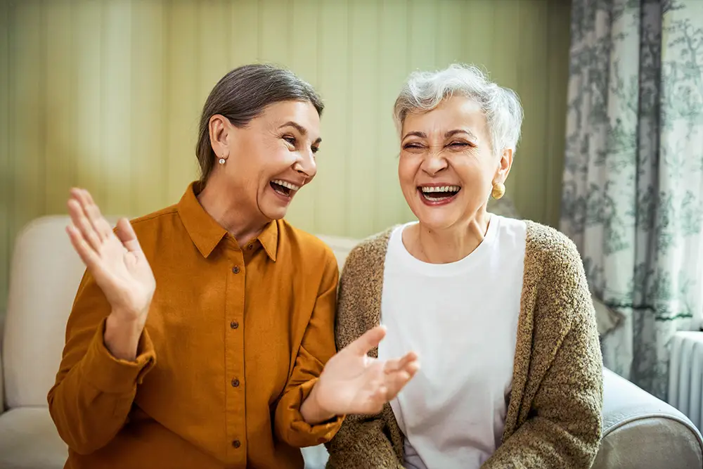 Two older women sitting on a couch, smiling and laughing together in a living room.