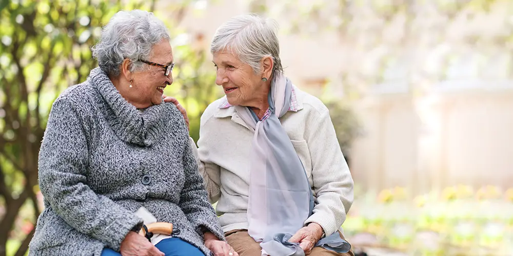 Two elderly women sit outdoors on a bench, smiling and talking to each other, with greenery and flowers in the background.