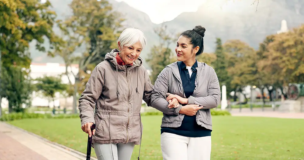 An elderly woman with a cane walks outside with a younger woman, who is holding her arm. They are both wearing jackets and walking in a park.