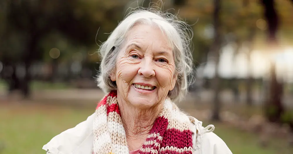 Elderly woman with gray hair smiling outdoors, wearing a cream jacket and a red and white striped scarf. Trees and blurred greenery are in the background.