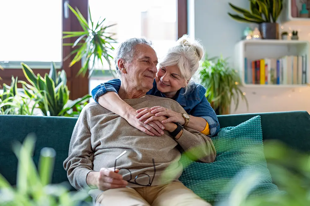 An older woman hugs an older man from behind as they sit together on a couch in a living room with houseplants and books in the background.