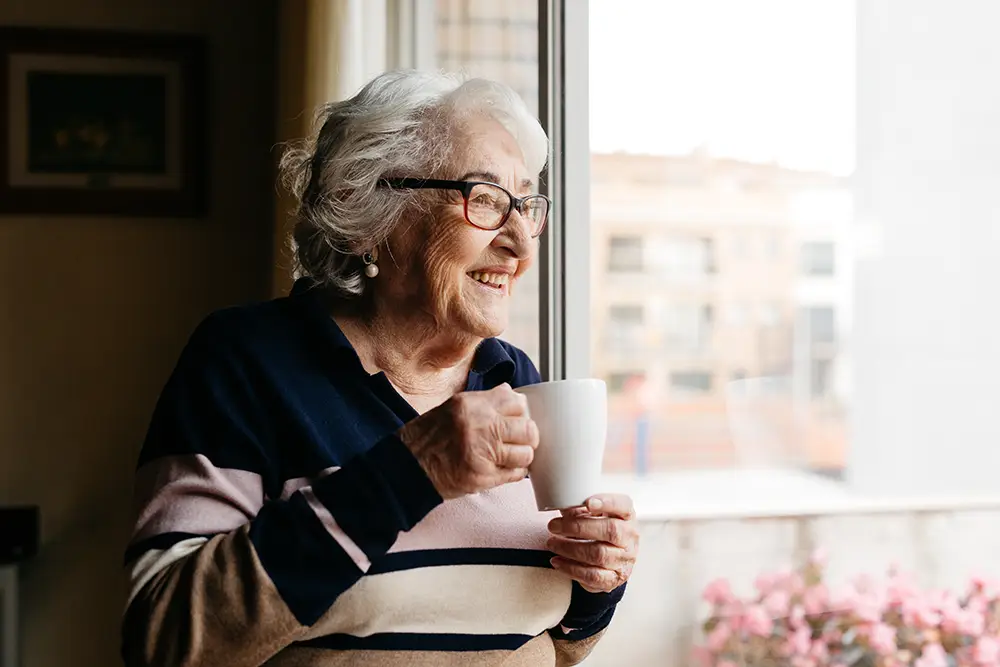 Older woman with glasses and gray hair holds a white mug and looks out a window, smiling, with buildings and pink flowers visible outside.