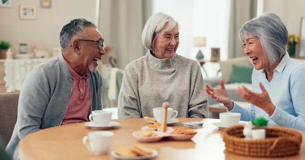 Three older adults sit around a table with coffee and snacks, engaged in conversation and smiling in a cozy indoor setting.