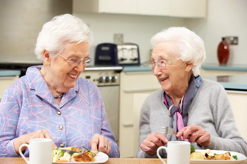 Two elderly women sit at a table in a kitchen, smiling and eating a meal together, with plates of food and mugs in front of them.