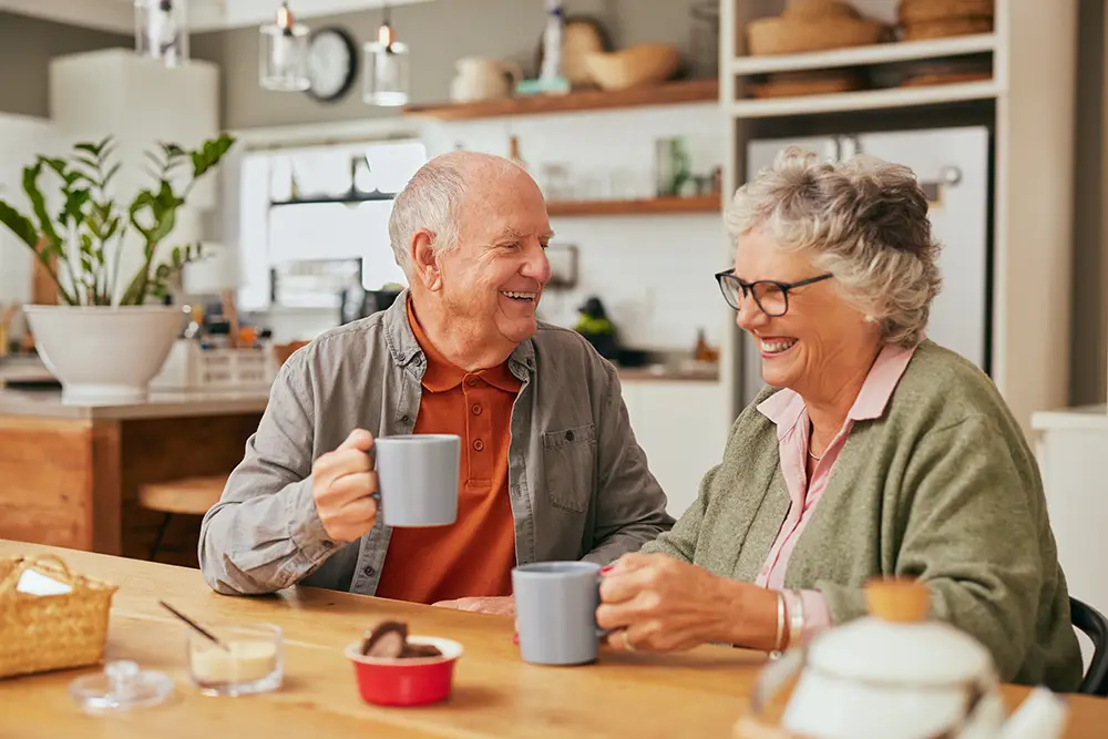 Two older adults sit at a kitchen table, smiling and holding mugs, with food and kitchen items visible in the background.