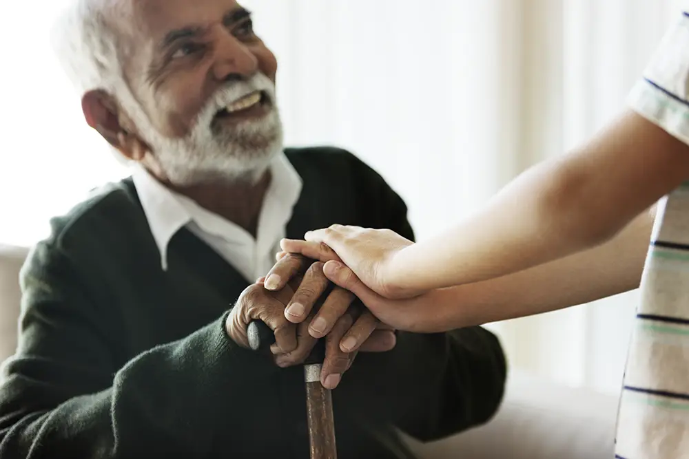 An elderly man with a beard holding a cane smiles as a younger person places their hands on his in a gesture of support.