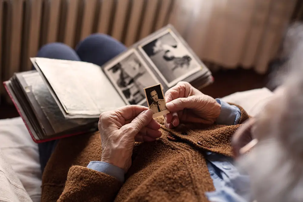 An elderly person sits holding an old black-and-white photo, with an open photo album on their lap.