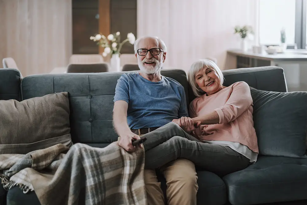 An older man and woman sit together on a couch, smiling and holding hands, with a blanket over their legs in a cozy living room.