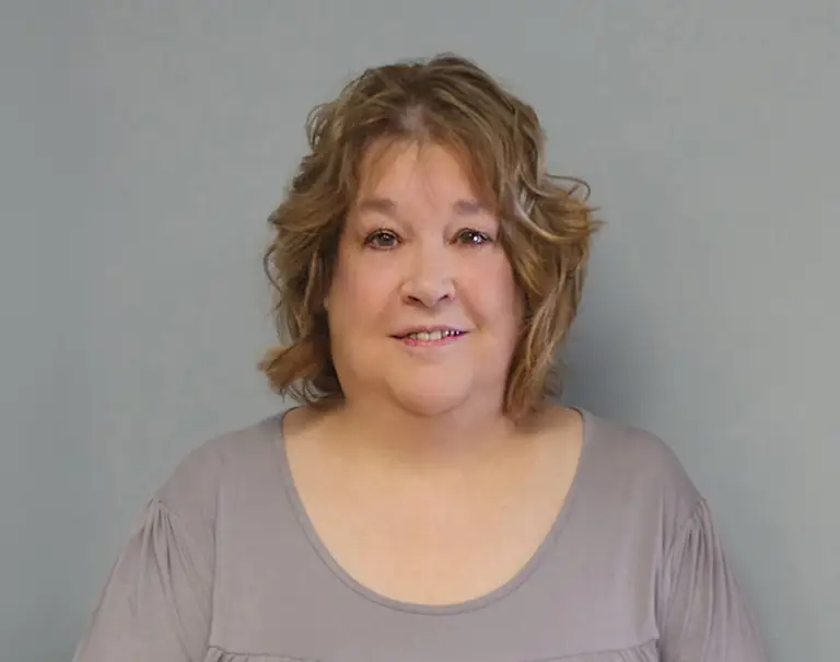 A woman with wavy, shoulder-length light brown hair smiles at the camera. She is wearing a light gray top and is posed against a plain gray background.