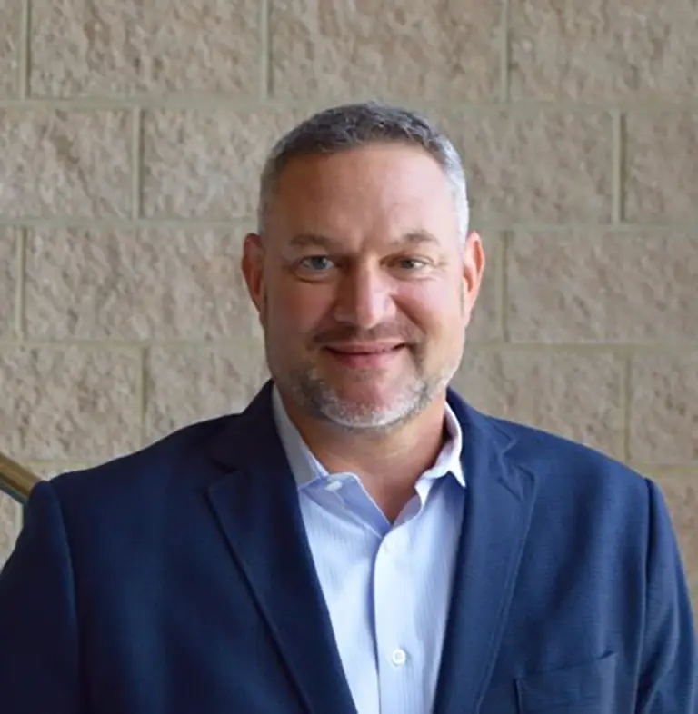 A man with short gray hair and a trimmed beard wearing a navy blazer and light blue shirt stands in front of a tan brick wall, smiling at the camera.