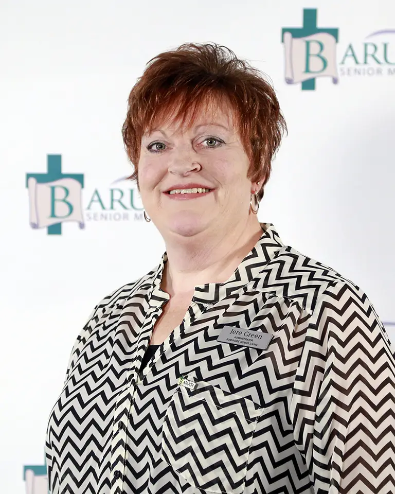 A woman with short red hair wearing a black and white zigzag patterned blouse and a name tag stands in front of a Baruch Senior Ministries backdrop.