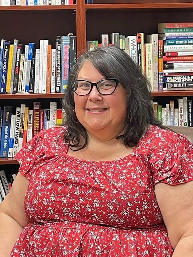 Woman with glasses and long dark hair, wearing a red floral blouse, sits in front of a bookshelf filled with various books.