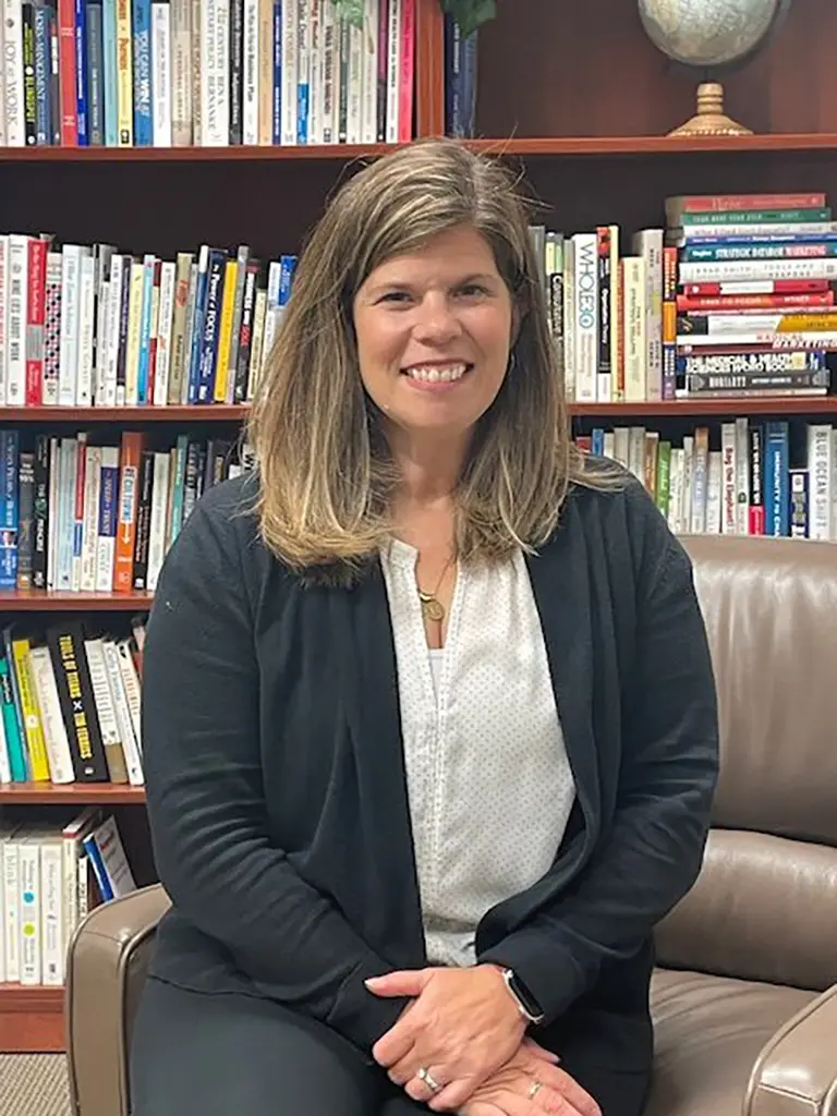 A woman with shoulder-length brown hair sits on a brown chair in front of a bookshelf filled with books, smiling at the camera.