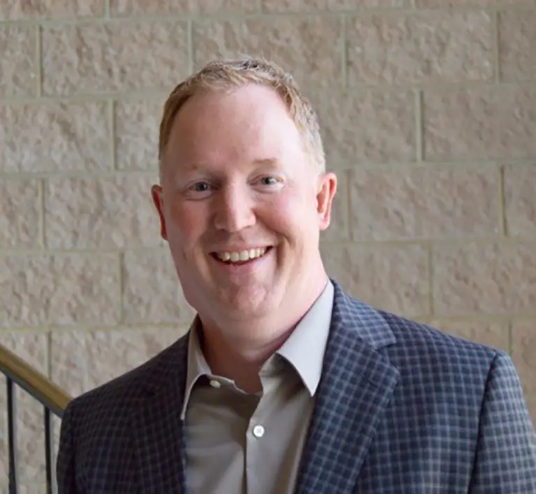 A man with short light hair wearing a checked blazer and light shirt stands in front of a beige brick wall, smiling at the camera.