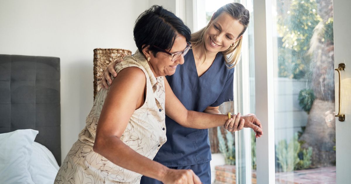 A caregiver in scrubs assists an older woman with short hair as she stands up near a bed in a sunlit room, highlighting the personal support found in in-home care vs assisted living.