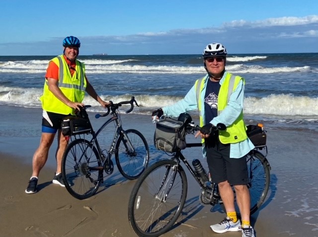 Two people wearing helmets and reflective vests stand with their bicycles on a sandy beach near the ocean, waves and blue sky in the background, taking part in the Biking for Baruch event.
