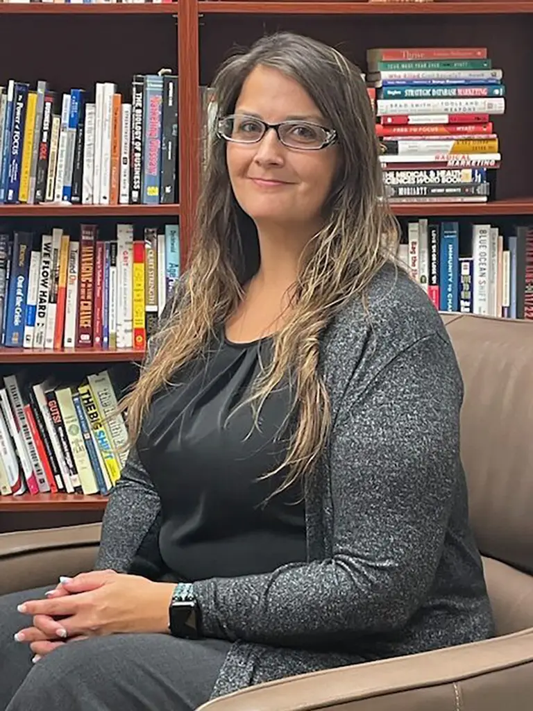 A woman with long brown hair and glasses sits in an armchair, hands folded, in front of a bookshelf filled with books.