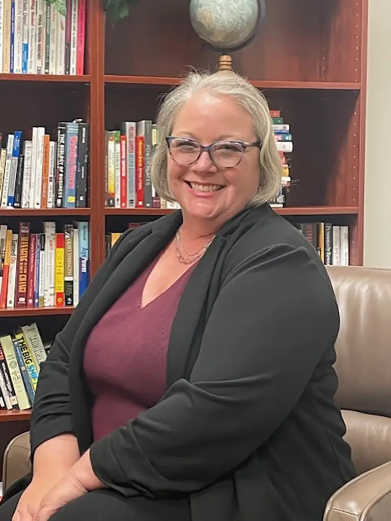 A woman with gray hair and glasses, wearing a black blazer and maroon top, is seated and smiling in front of a bookshelf filled with books.
