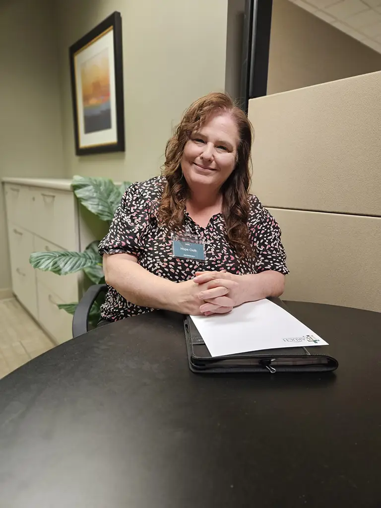A woman with long brown hair sits at a round table in an office, smiling, with her hands resting on a closed folder and a notepad in front of her.