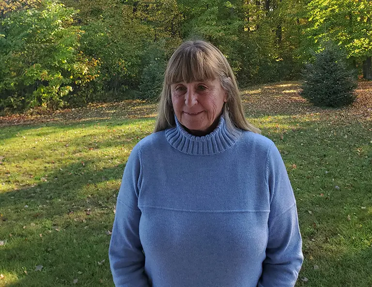 A woman with long hair and bangs, wearing a light blue sweater, stands outside on a grassy area with trees and sunlight in the background.