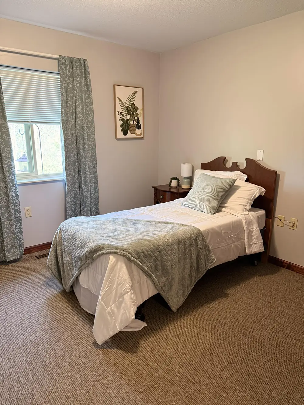 HorizonClare-1 A neatly made twin bed with gray bedding and pillows is next to a window with patterned curtains in a simple, carpeted bedroom. A small side table holds a lamp and decor.
