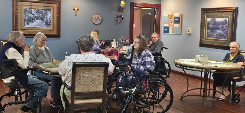 HorizonClare-4 Several elderly people sit in wheelchairs and chairs around tables in a care facility room with framed artwork on the walls.
