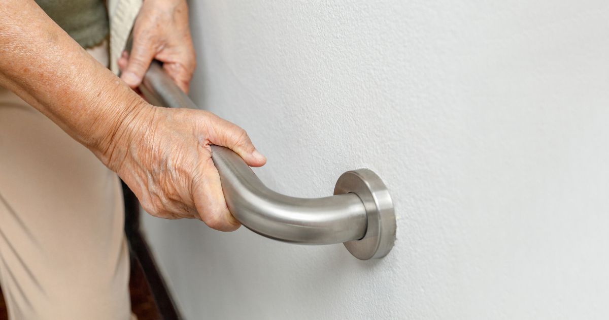 Close-up of an elderly person’s hands holding a metal grab bar mounted on a wall, highlighting aging in place safety concerns and the importance of a senior home safety checklist.