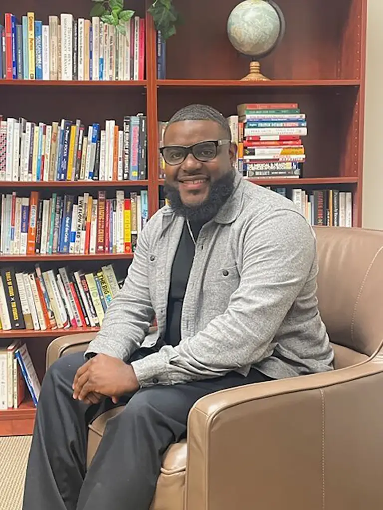 A man with a beard and glasses sits in a brown armchair, smiling, with a bookshelf filled with books and a globe behind him.