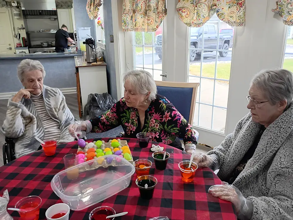 HydePark-4 Three elderly women sit at a table with plastic cups of colored dye, dyeing Easter eggs together indoors. A man works in the background near a kitchen area.