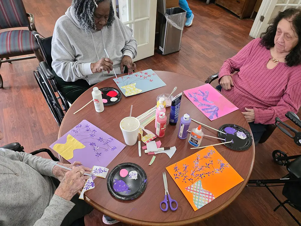 HydePark-6 Three women sit around a table doing arts and crafts, painting and gluing decorative paper onto colored sheets. Art supplies and finished pieces are scattered on the table.