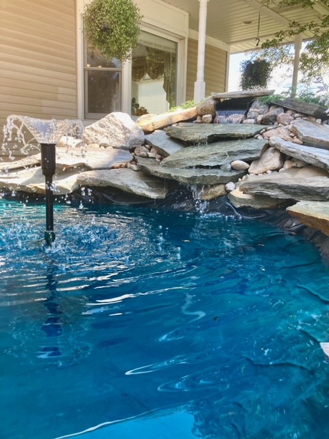 A backyard pond with clear blue water, a small waterfall over stones, and water splashing from a fountain near a house patio—Dream It, Believe It, Achieve It in your own outdoor oasis.