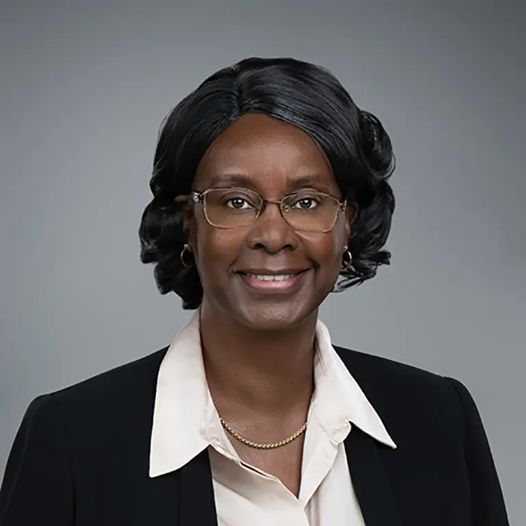 A woman with glasses, wearing a black blazer, cream blouse, and gold jewelry, smiles at the camera against a plain gray background.