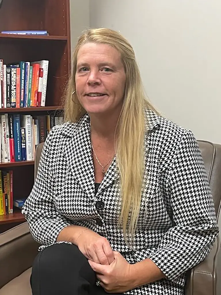 A woman with long blonde hair sits in an office chair, wearing a black and white houndstooth jacket, with a bookshelf filled with books in the background.