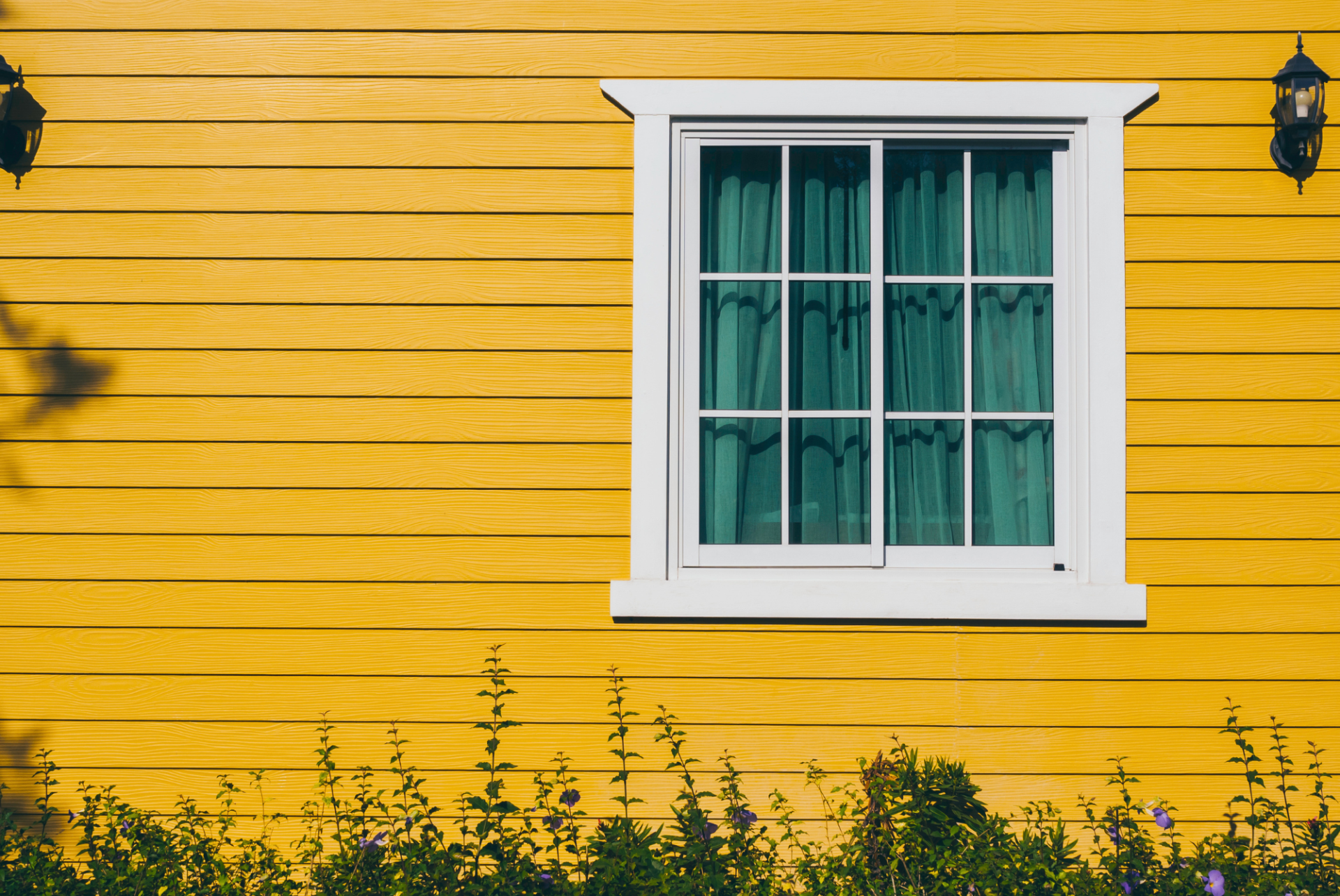 A white-trimmed window with closed curtains is set in the middle of a bright yellow wooden house wall, where plants grow at the base—showcasing how making a difference starts with every thoughtful detail.