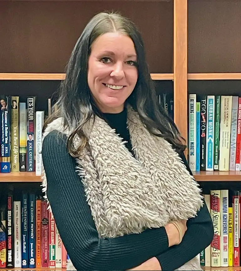 A woman with long brown hair, wearing a black top and a light furry vest, stands in front of a bookshelf filled with books, smiling with arms folded.