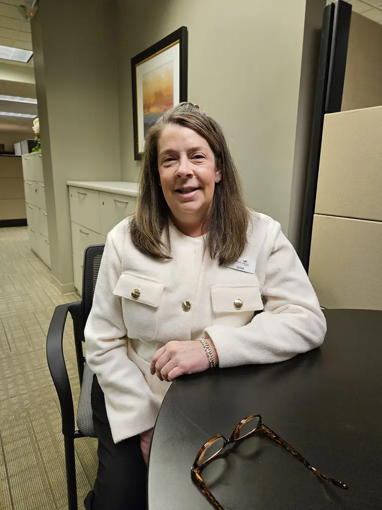 A woman with long brown hair wearing a cream jacket sits at a table in an office, with eyeglasses on the table and filing cabinets in the background.