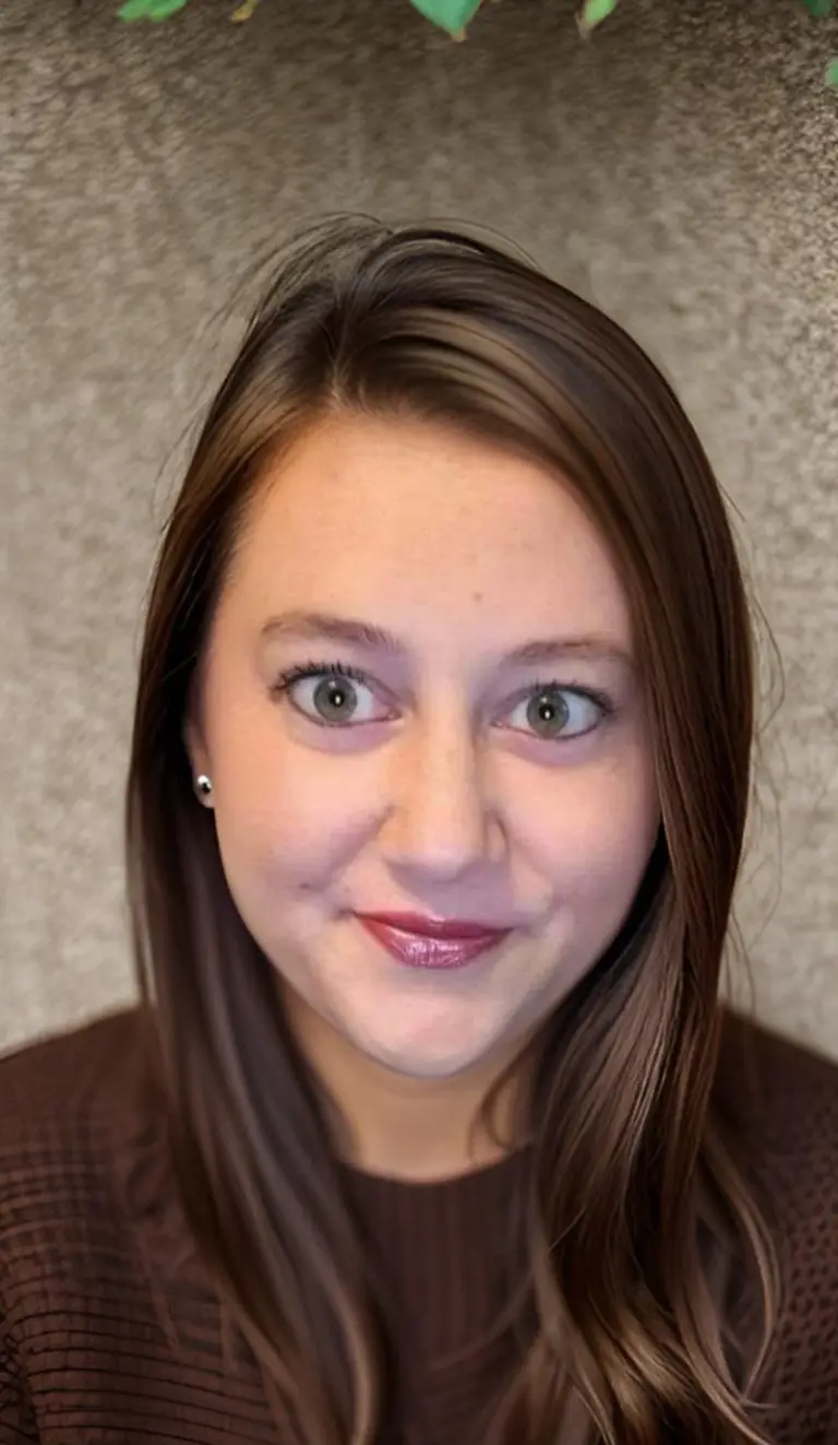 A woman with straight brown hair and green eyes, wearing a brown top, is smiling slightly at the camera against a textured beige background.