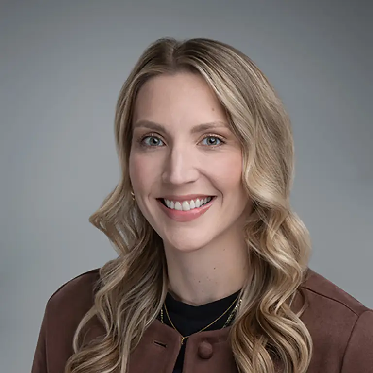 Smiling woman with long blonde wavy hair wearing a brown jacket over a black top, posing in front of a neutral gray background.