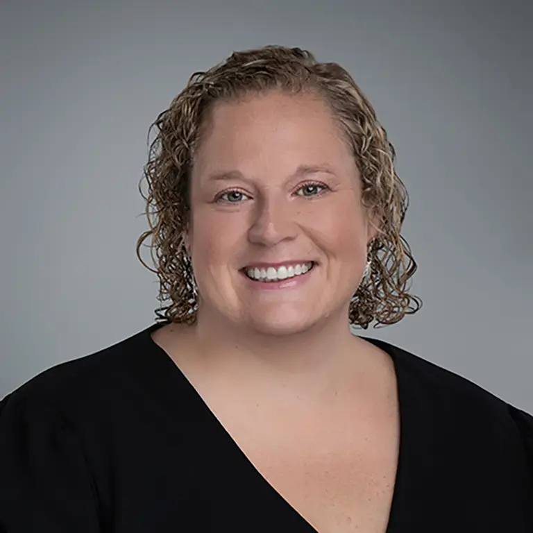 Smiling woman with short curly blonde hair wearing a black top, posed against a plain gray background.