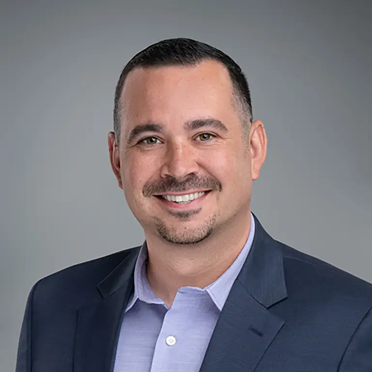 Man with short dark hair, trimmed beard, and mustache wearing a navy suit jacket and light purple shirt, smiling at the camera with a neutral gray background.