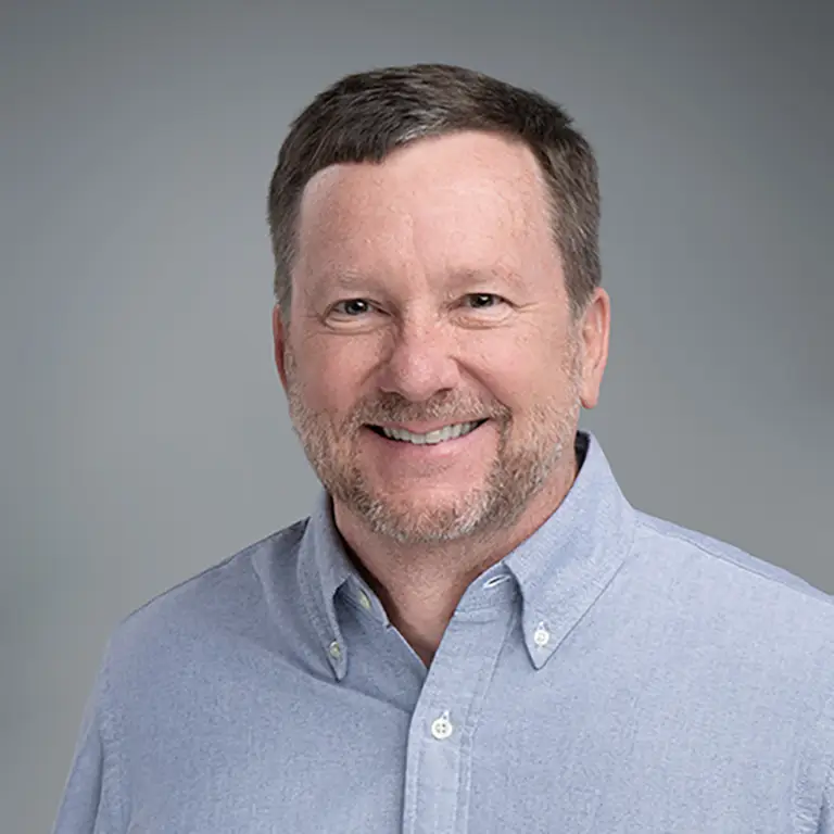 A man with short brown hair and a beard, wearing a light blue button-down shirt, smiles at the camera against a plain gray background.