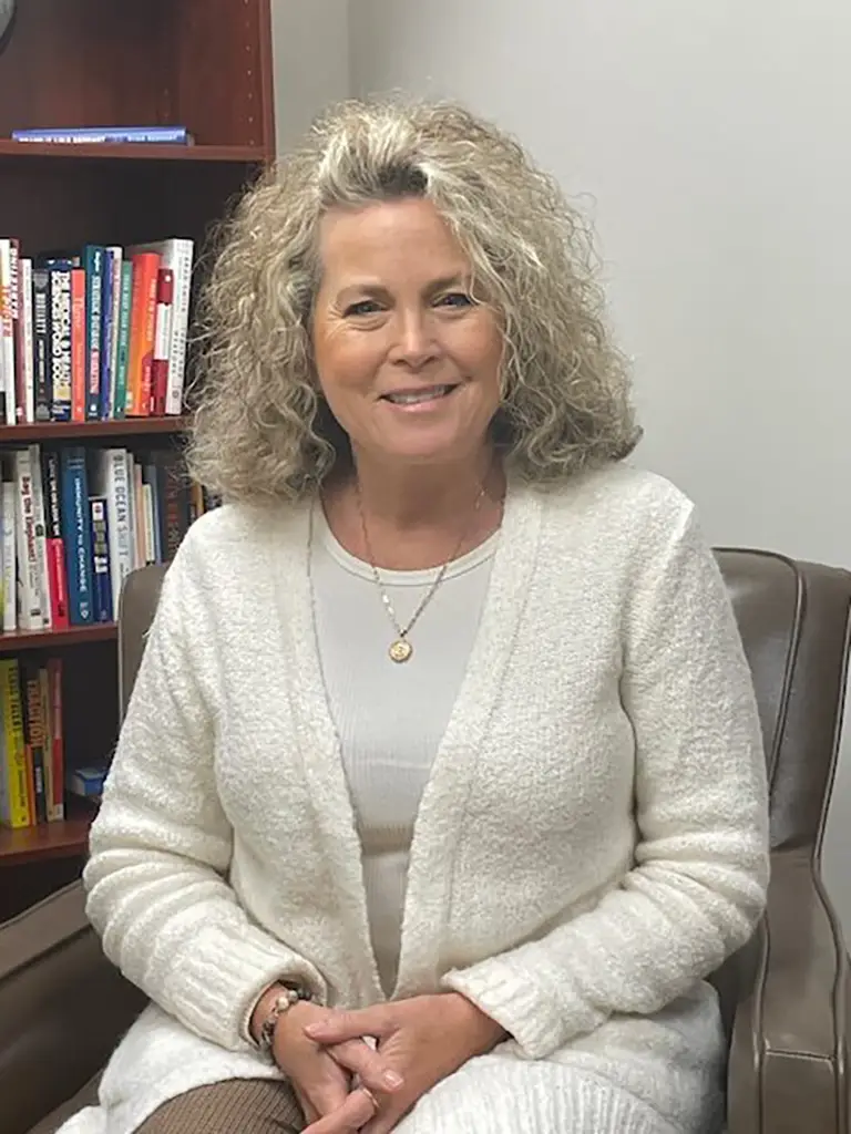 A woman with curly blonde hair sits in an armchair, smiling, wearing a light-colored sweater. A bookshelf filled with books is in the background.