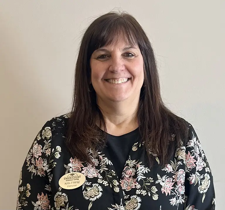 A woman with long brown hair and a floral-patterned top smiles at the camera. She is wearing a name badge.