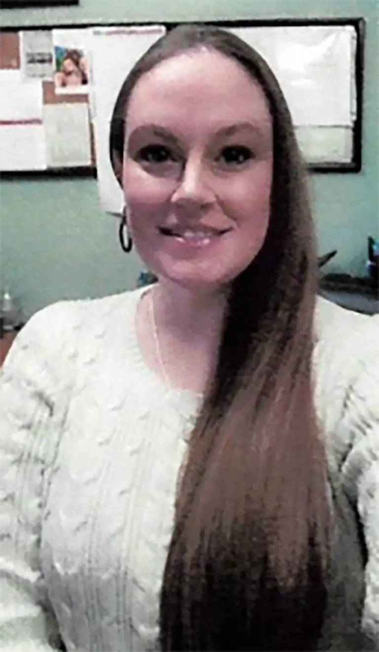 A woman with long straight hair, wearing a light-colored sweater, sits indoors with bulletin boards and papers visible in the background.