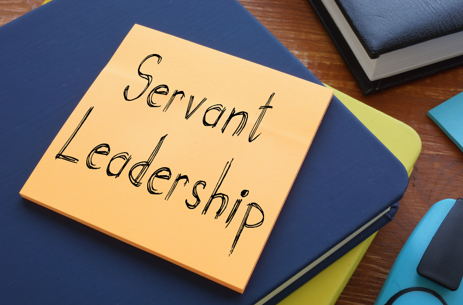 A sticky note with "Servant Leadership" written on it is placed on top of stacked books and folders on a wooden desk, highlighting the Gift of Administration in Spiritual Care.