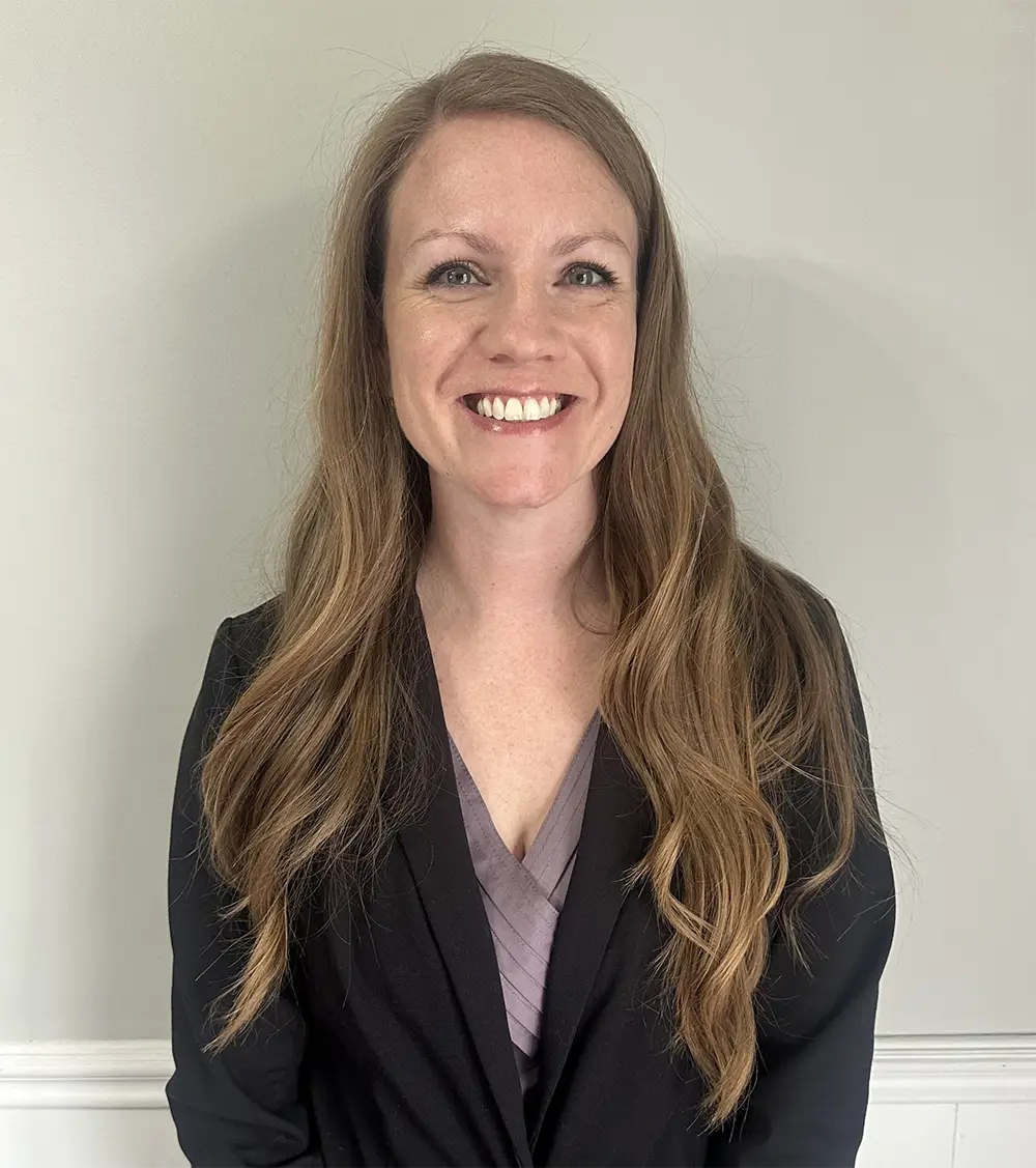 A woman with long, wavy brown hair wearing a black blazer and light purple top stands smiling in front of a plain light-colored wall. A woman with long, wavy brown hair wearing a black blazer and light purple top stands smiling in front of a plain light-colored wall.