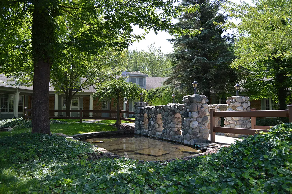 A small stone pond and wall in front of a building, surrounded by trees, ivy, and a wooden fence on a sunny day. A small stone pond and wall in front of a building, surrounded by trees, ivy, and a wooden fence on a sunny day.